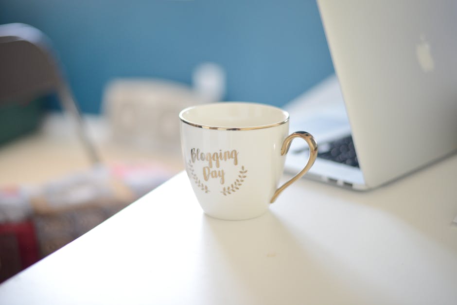 Close-up of a ceramic coffee cup labeled 'Blogging Day' on a white desk beside a laptop at a home office.