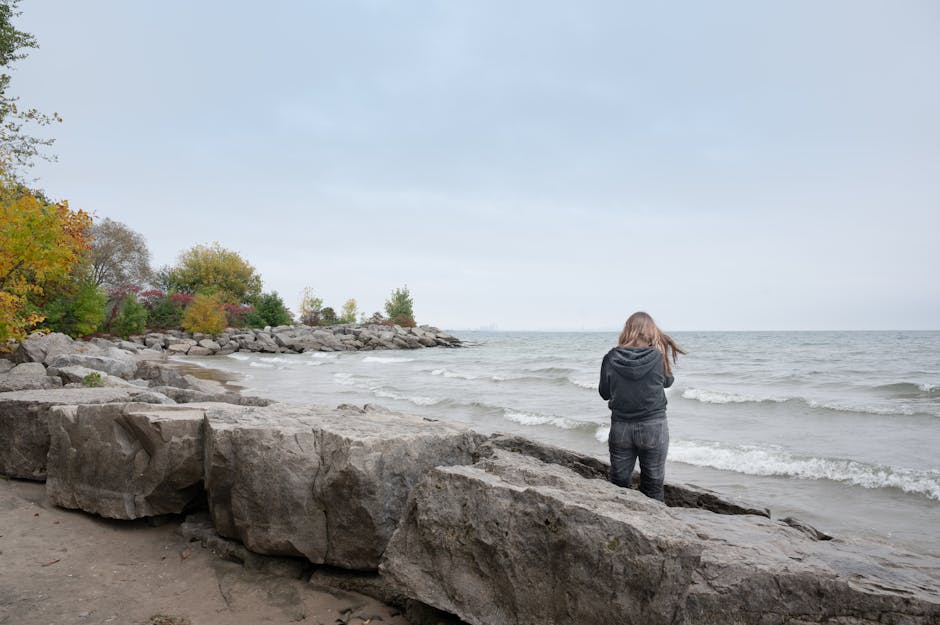 A woman stands alone by the rocky shore of a lake in Mississauga, ON, Canada, amidst the autumn scenery.