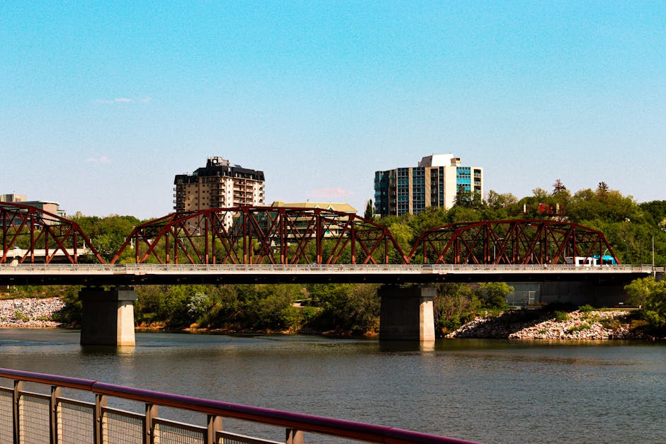 Urban bridge crossing river with backdrop of city buildings under clear blue sky.